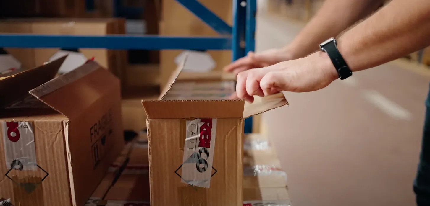 Hands opening a taped cardboard box labeled with fragile symbol on a warehouse shelf.
