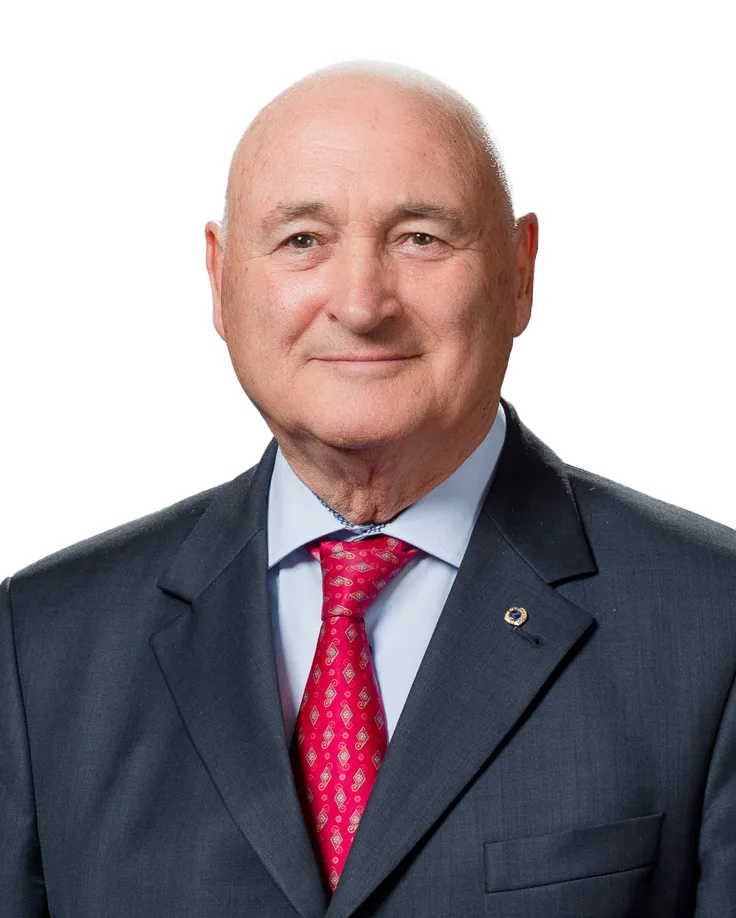 Smiling bald man in a dark suit, light blue shirt, and red patterned tie against a white background.
