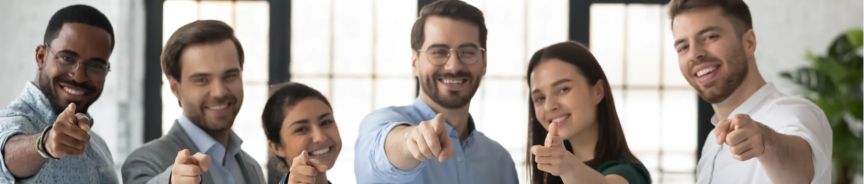 Diverse group of six smiling young professionals pointing forward in an office setting.