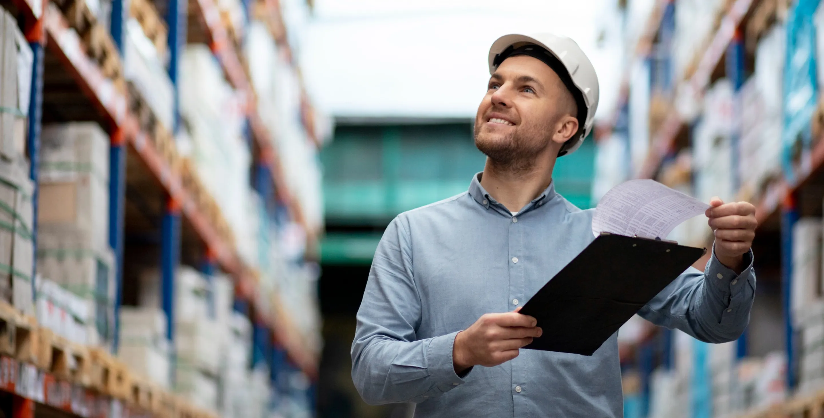 Man wearing a white safety helmet and blue shirt holding a clipboard inside a warehouse.