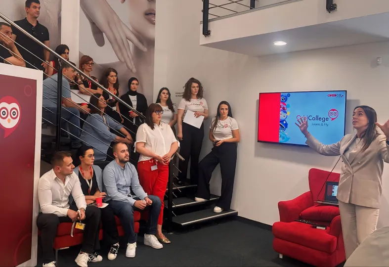 A woman giving a presentation to a group seated on stairs and couches in a modern office, with a screen displaying 'Orbi College Learn & Fly'.