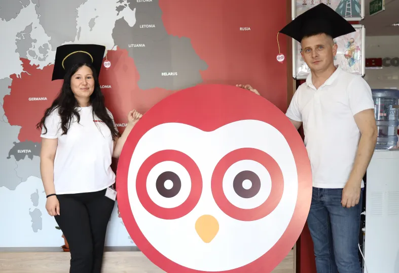 A woman and a man wearing black graduation caps stand on either side of a large red and white owl logo, with a map of Europe in the background.