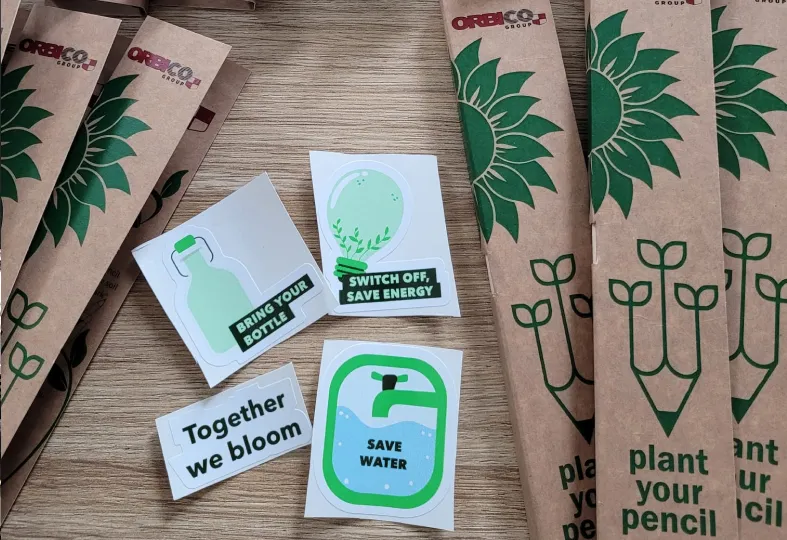 Stickers with environmental messages like ‘Bring your bottle,’ ‘Switch off, save energy,’ ‘Together we bloom,’ and ‘Save water’ next to plantable pencils in cardboard sleeves.