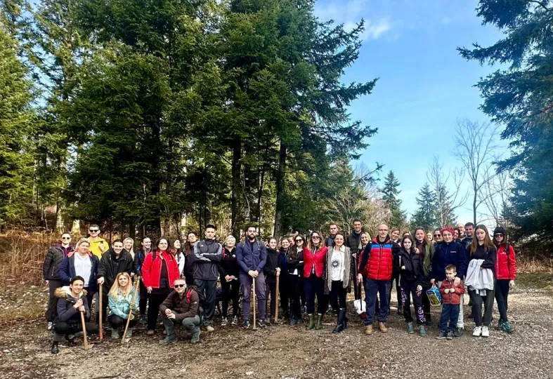 Group of people posing outdoors in a wooded area on a sunny day, some holding walking sticks.