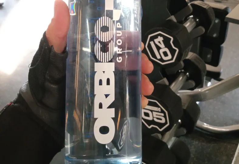 Hand wearing a black glove holding a clear water bottle with ORBICO GROUP branding, with gym dumbbells in the background.