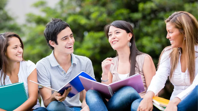 Group of four young adults sitting outdoors, smiling, and holding colorful notebooks while engaging in conversation.