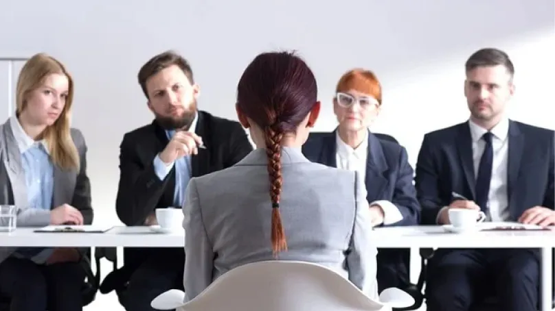 A woman with braided hair facing a panel of four businesspeople in a job interview setting.