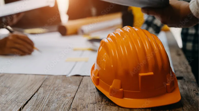 Orange construction hard hat on a wooden table with blurred hands working on blueprints in the background.