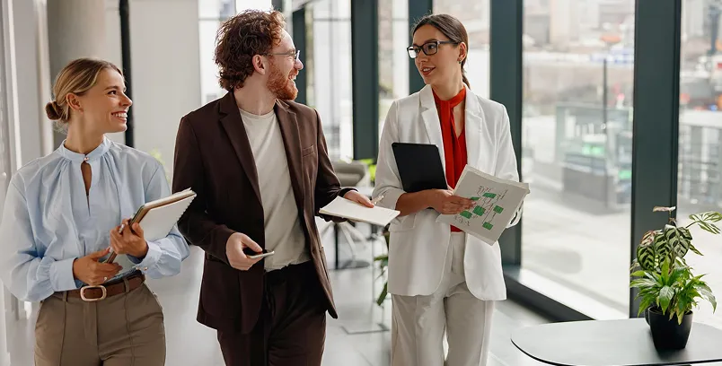 Three business professionals walking and discussing documents in a modern office with large windows.