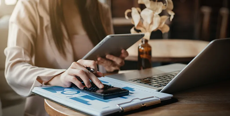 Person using a calculator and holding a tablet near an open laptop and clipboard with charts on a wooden table.