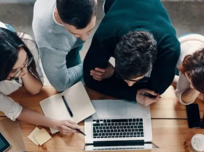 Four people gathered around a laptop on a wooden table, one pointing at the screen with a pen, with notebooks, a smartphone, and a coffee cup nearby.