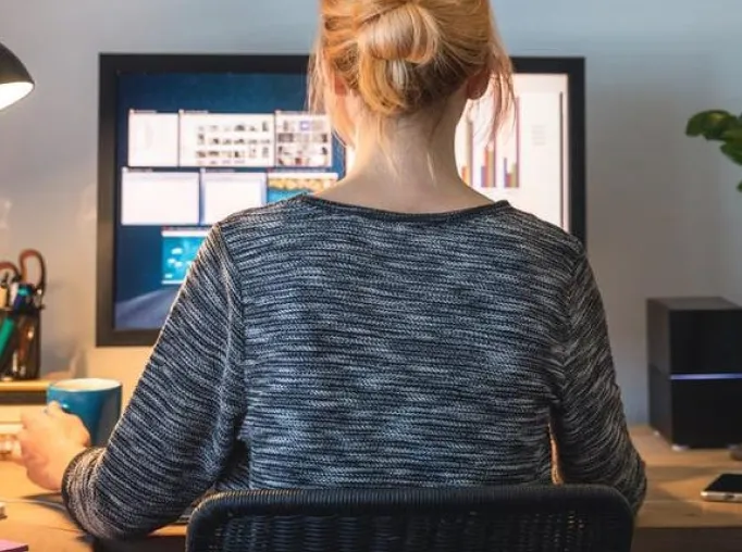 Person with blonde hair in a bun sitting at a desk holding a blue mug and looking at a computer monitor displaying multiple open windows.