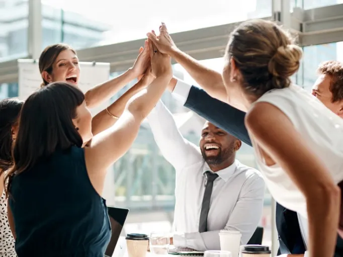 Diverse group of business colleagues smiling and giving a group high-five around a table in a modern office.