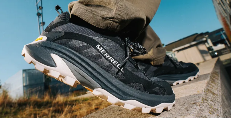 Close-up of a person wearing black Merrell hiking shoes standing on a stone ledge outdoors with clear blue sky.