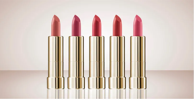 Five gold-cased lipsticks in varying shades of pink and red arranged in a row on a reflective surface.