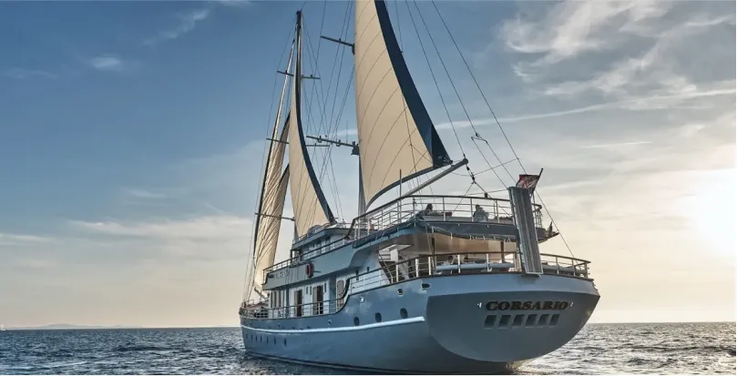 Large white sailing ship named Corsario with raised sails on calm ocean waters under a partly cloudy sky at sunset.