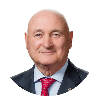 Portrait of a bald older man in a black suit, blue shirt, and red patterned tie against a white background.