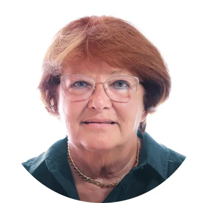 Portrait of an older woman with short reddish hair and glasses wearing a dark collared shirt and gold necklace.