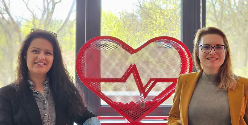 Two smiling women sitting indoors with a large red wire-frame heart and heartbeat symbol between them in front of a window.