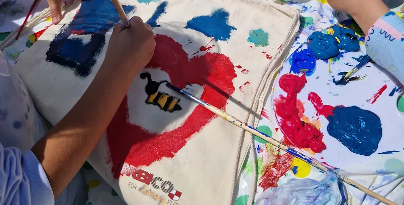 Person painting a bee inside a red heart on a canvas tote bag with various paint colors on a palette.