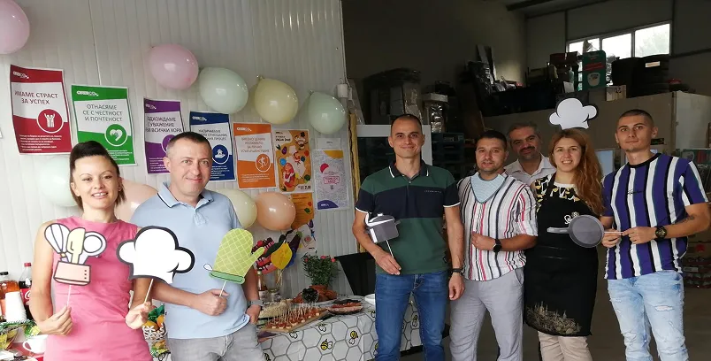 Group of seven people in a room decorated with balloons and colorful posters, holding kitchen-themed photo props and smiling.